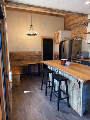 A rustic kitchen with a wooden bar, stools, a refrigerator, and a coffee machine.