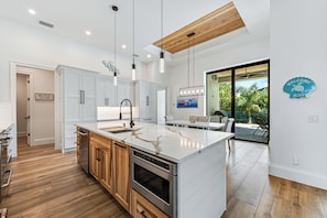 A modern kitchen with white cabinetry, a large island, stainless steel appliances, pendant lighting, and a view of the outdoors. - Luxury Coastal Beach Escape w/ Pool, Hot Tub & walkable to the beach (Marco Island)