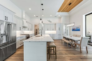 A modern kitchen with white cabinetry, stainless steel appliances, a central island, and a dining area with wooden flooring and a bench. - Luxury Coastal Beach Escape w/ Pool, Hot Tub & walkable to the beach (Marco Island)