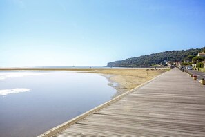 Ein hölzerner Steg führt zu einem Strand mit ruhiger See, klarem Himmel und einer Hügelside mit Gebäuden.