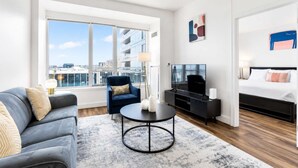 Una habitación de hotel moderna con un ventanal grande, un sofá, una silla, un televisor, una cama y una mesita.