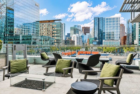 Eine Dachterrasse mit Sitzgelegenheiten im Freien, Blick auf eine Stadtlandschaft und einer Glasbrüstung.