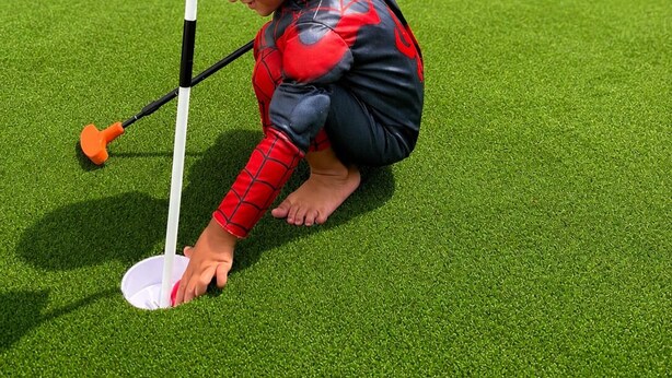 Een kind in een Spider-Man kostuum aan het golfen op kunstgras.