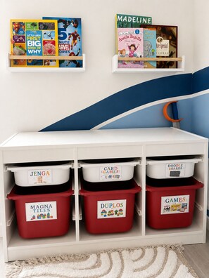 A children's bookshelf with books and storage bins labeled for games and activities. - Key and Code Stays: The Adventure House (St. Augustine)