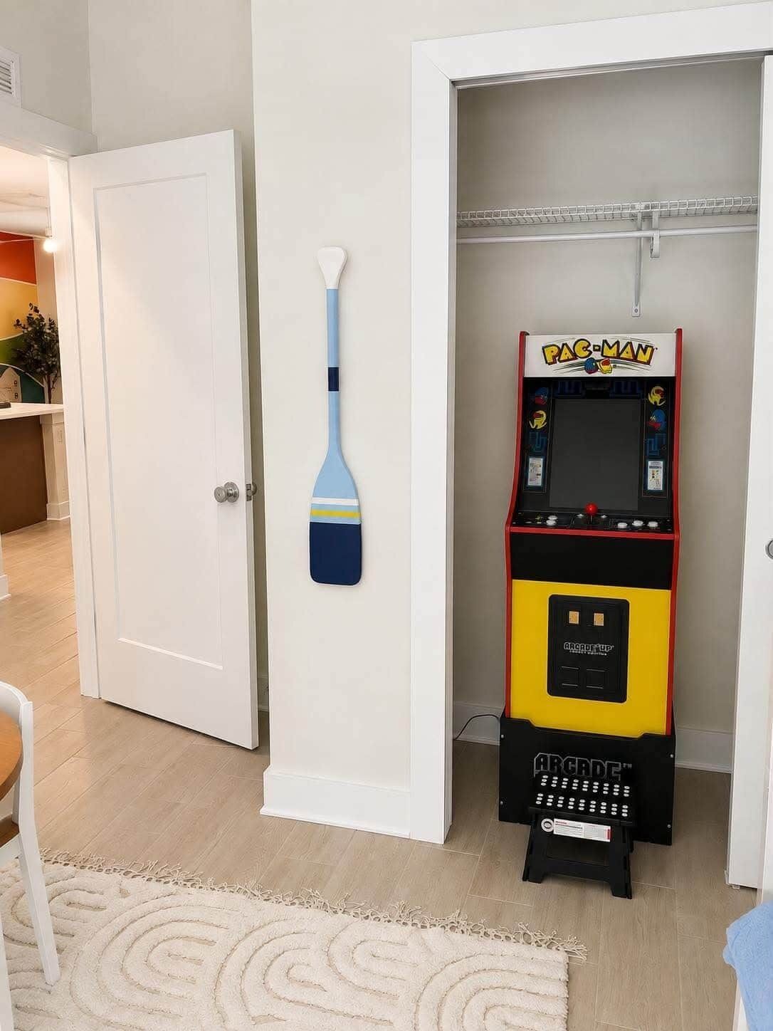 Uma máquina de arcade Pac-Man em um corredor com uma porta branca e um remo fixado na parede.