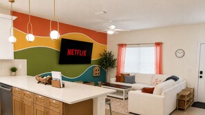 Een moderne woonruimte met een kookeiland, een witte bank, een televisie waarop 'NETFLIX' wordt weergegeven en een plafondventilator.