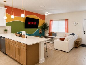 A modern living space with a kitchen island, a white sofa, a television displaying 'NETFLIX', and a ceiling fan. - Key and Code Stays: The Adventure House (St. Augustine)