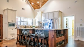 A kitchen with white cabinets, a stone island with stools, a stainless steel refrigerator, and a wooden ceiling.