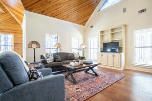 A spacious living room with a high ceiling, wooden beams, a large window, a television, and a bookshelf. - The Perfect Oxford Basecamp for Families, Friends & Rebels (Water Valley)