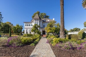 Un'ampia casa a due piani con un ingresso centrale, circondata da un giardino curato con diverse piante e un vialetto lastricato che conduce all'ingresso.