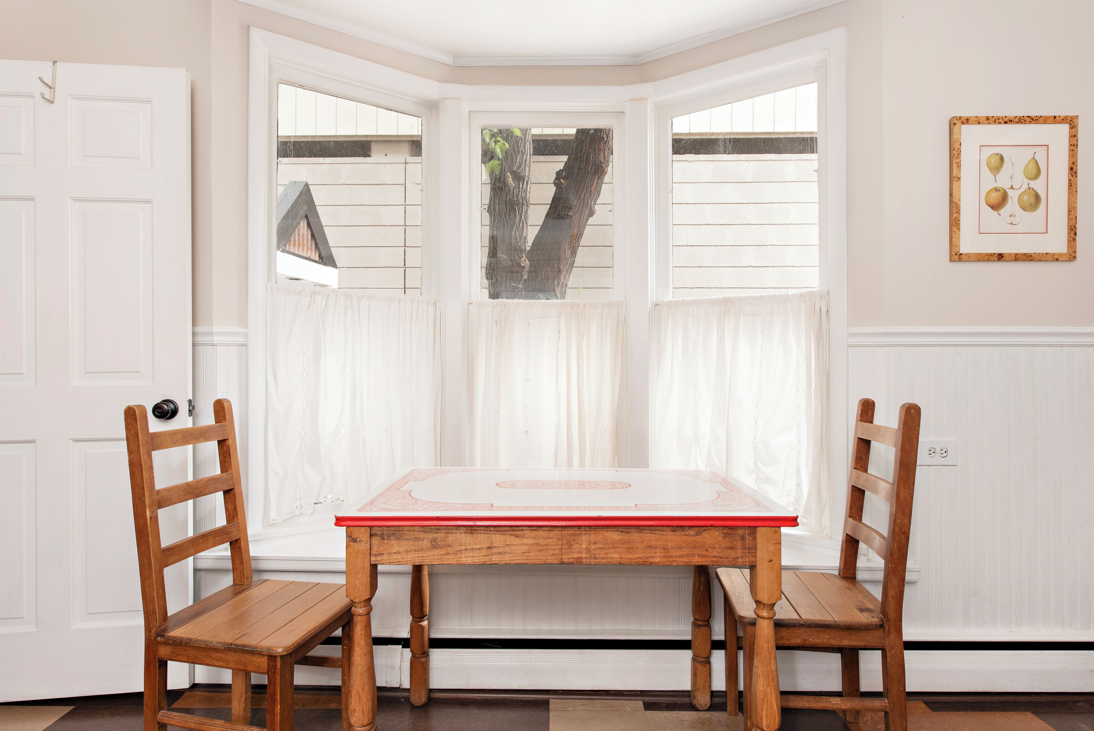 A room with a bay window, two wooden chairs, a table, and a framed picture on the wall.