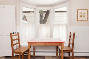 Ein Zimmer mit Erkerfenster, zwei Holzstühlen, einem Tisch und einem gerahmten Bild an der Wand.
