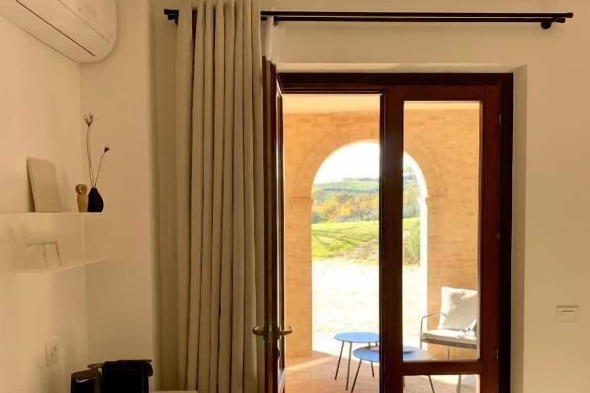 Una habitación con una puerta corredera que da a un espacio exterior, un escritorio blanco, una silla y un control remoto en el suelo.
