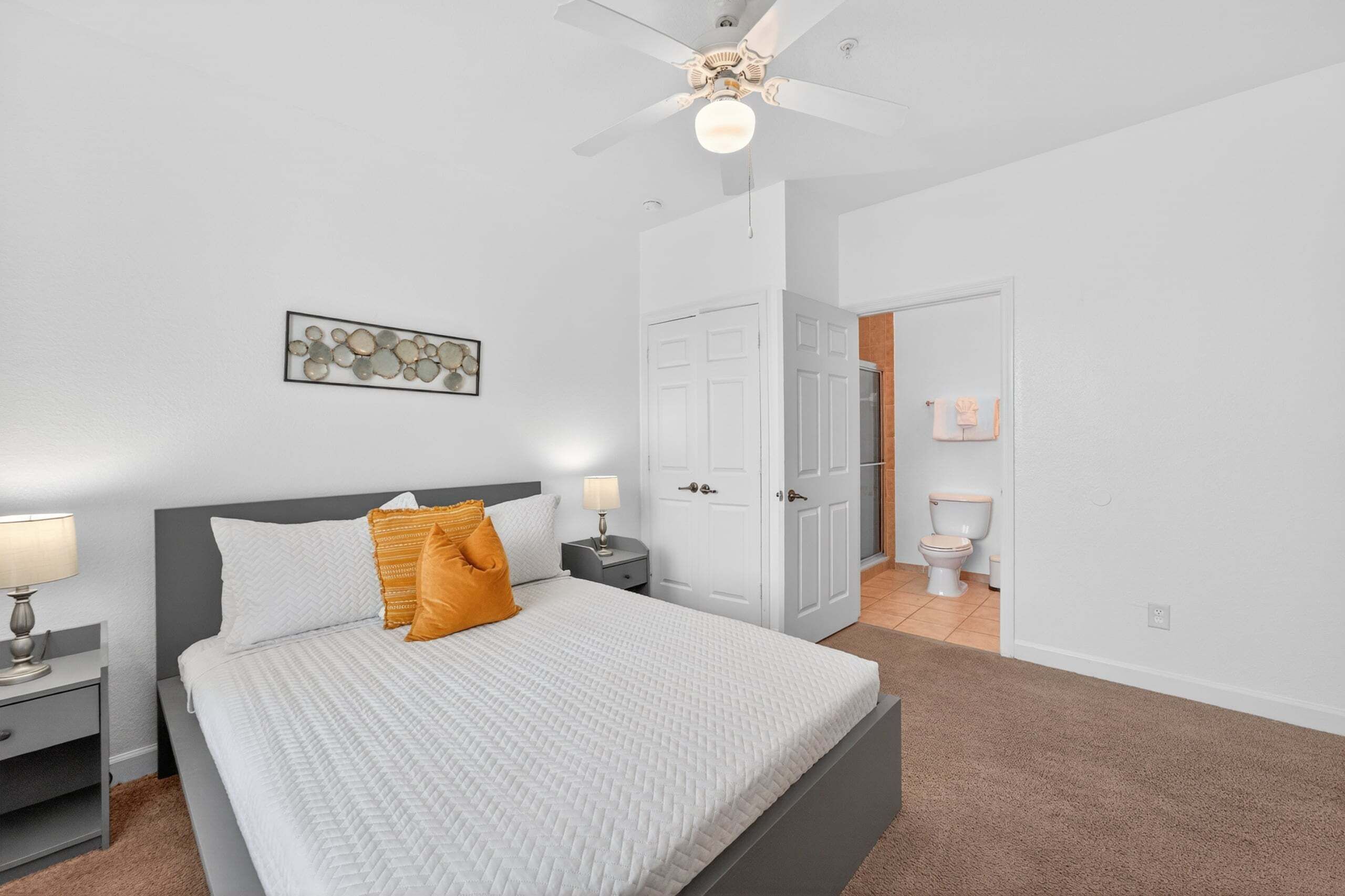 A bedroom with a bed, nightstand, lamp, ceiling fan, and a door leading to a bathroom.