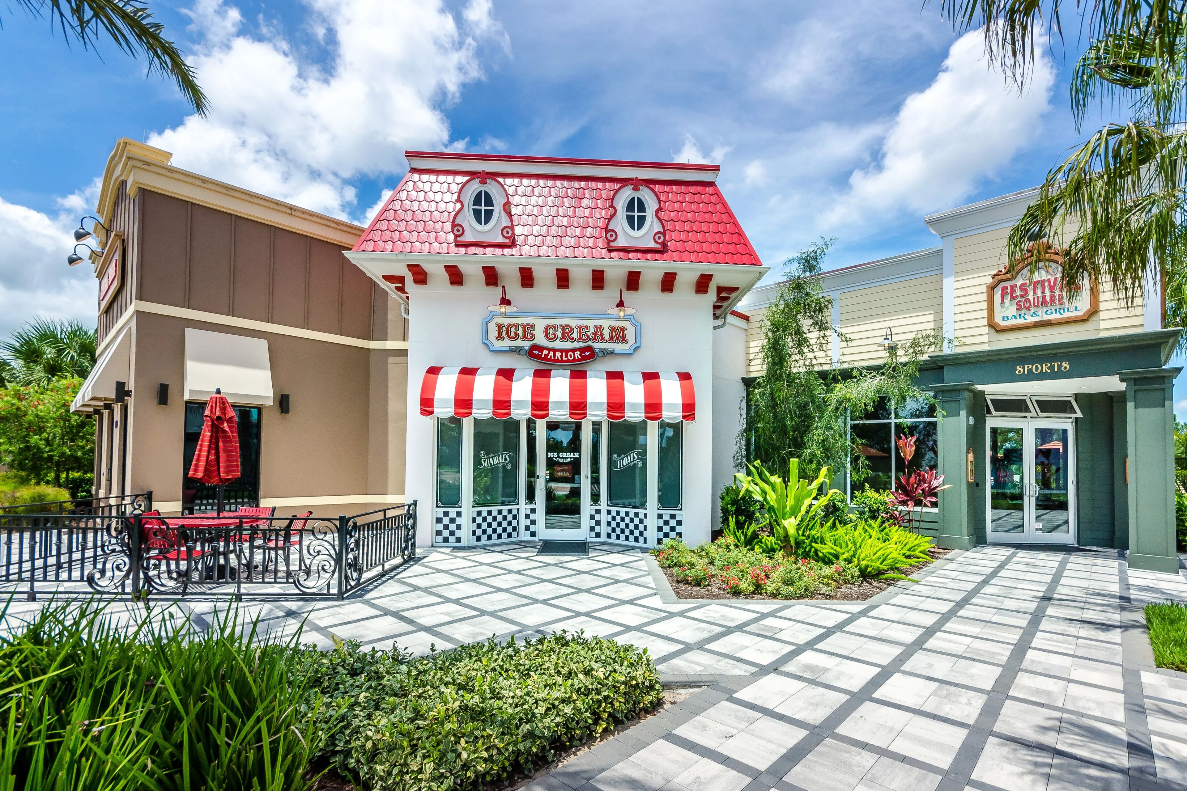 An ice cream parlor with a red and white striped awning, a checkered entrance, and outdoor seating with red chairs and a table.