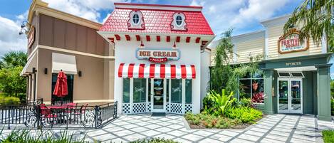 An ice cream parlor with a red and white striped awning, a checkered entrance, and outdoor seating with red chairs and a table.