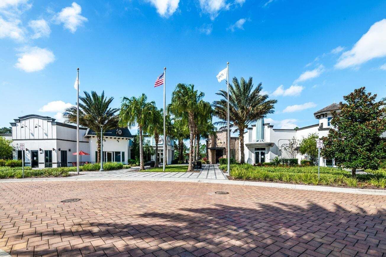 Una plaza pavimentada con palmeras, un asta con la bandera estadounidense y un edificio de fachada blanca.
