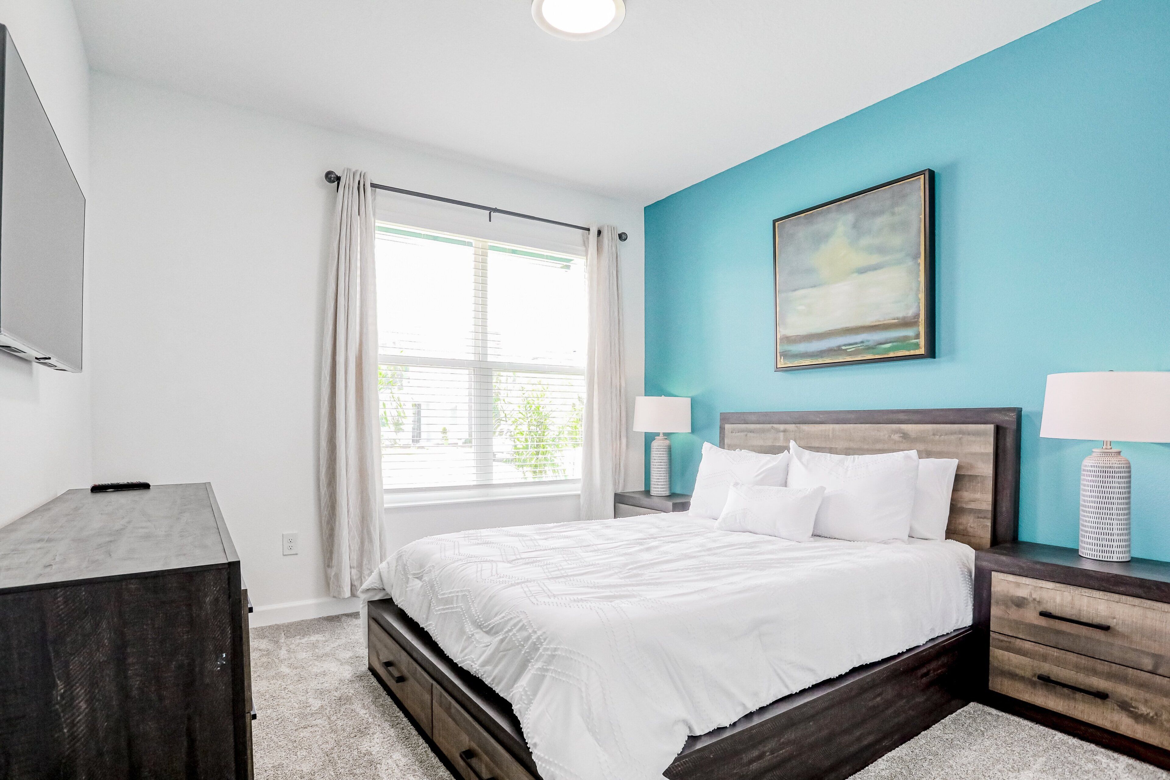 Une chambre à coucher comprenant un lit, des tables de chevet, une commode, une fenêtre avec des rideaux et un tableau au mur.