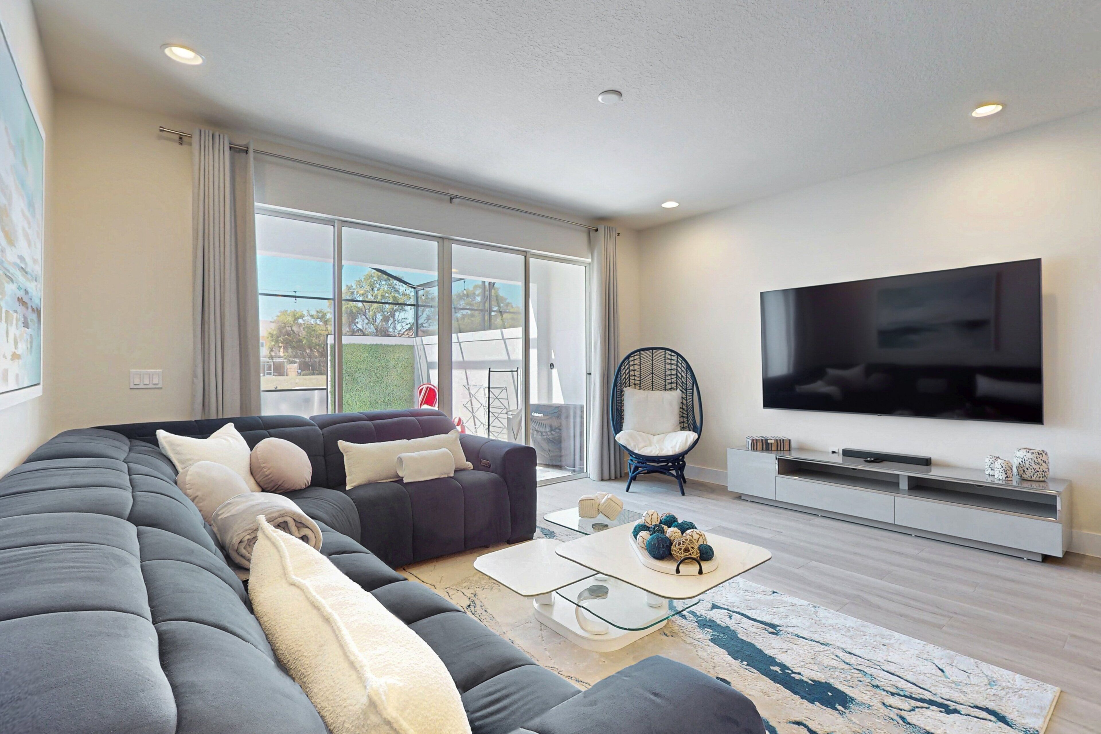 A modern living room with a sectional sofa, a flat-screen TV, a glass coffee table, and a large window.