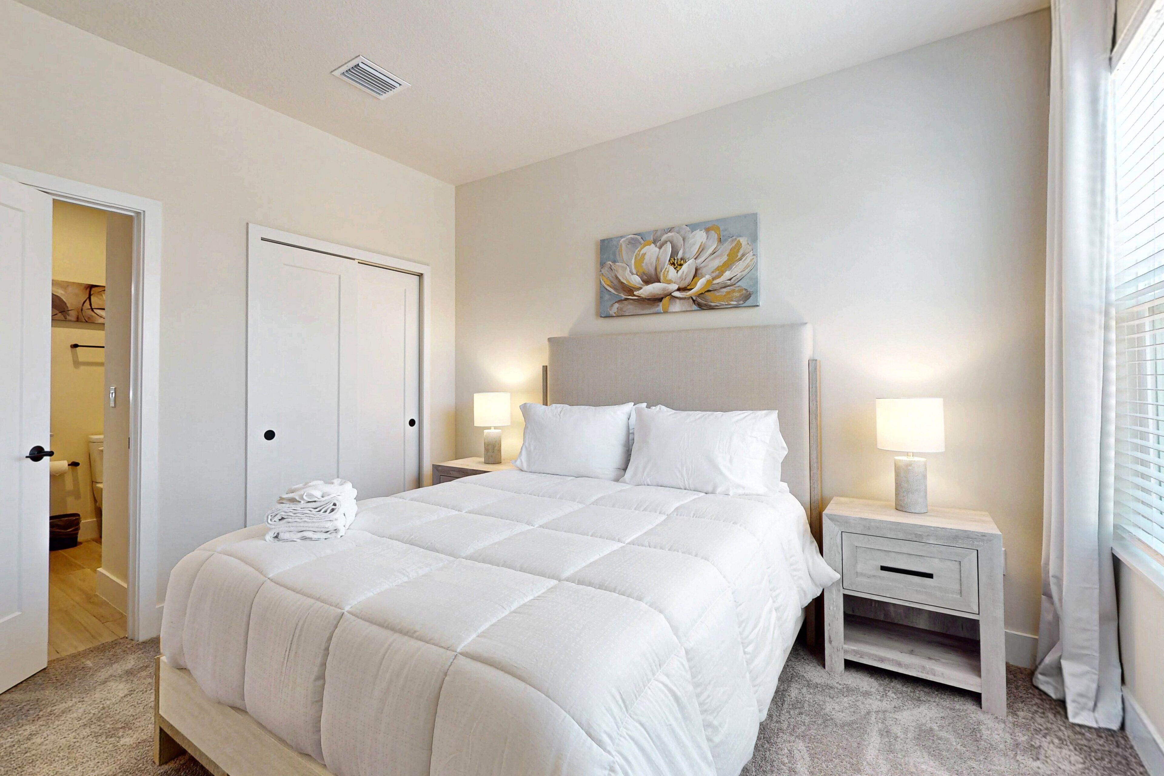A bedroom with a large bed, two bedside tables with lamps, a door, and a wall-mounted artwork.