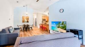 Ein Wohnzimmer mit Sofa, einem Esstisch mit Stühlen, einer Küchenzeile und einem Fernseher, der eine Strandansicht zeigt.