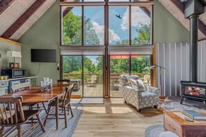 A spacious room with a large sliding glass door leading to an outdoor deck, a wooden dining table, and a fireplace. - Timber Ridge Cabin - Wifi | Fire pit | Horseshoe Pit | Outdoor Shower | Deck | Games (Berkeley Springs)