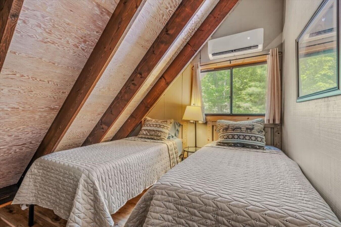 Een kamer met een schuine plafond, twee bedden met quiltdekens, een raam met uitzicht op bomen, en een airconditioning.