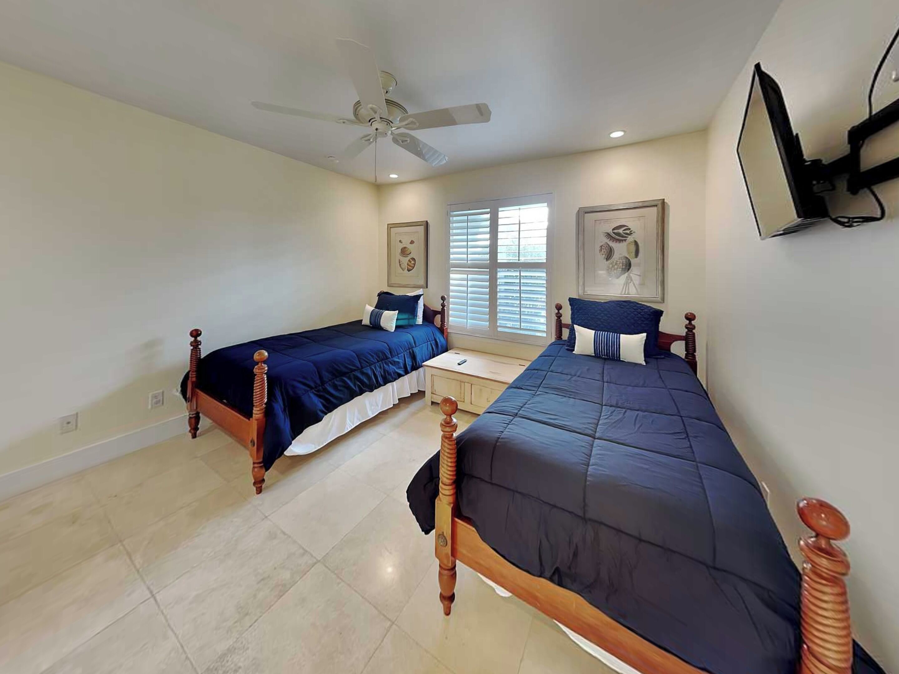 A bedroom with two single beds, a ceiling fan, a window with blinds, and a wall-mounted TV.
