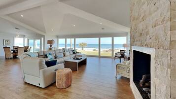 Uma sala de estar ampla com uma grande janela com vista para a praia, lareira e área de jantar com móveis de vime.