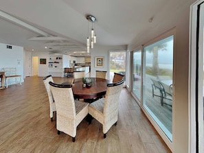 A spacious dining area with a round table and wicker chairs, a kitchen in the background, and large sliding glass doors leading to an outdoor patio. - Ocean Sunrise (Ponte Vedra Beach)