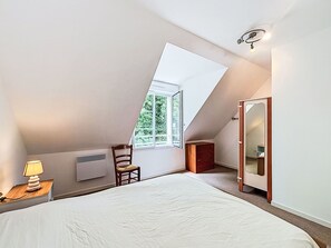 Ein Schlafzimmer mit schrägen Wänden, einem Bett, einem Sessel, einem Nachttisch, einer Kommode und einem Fenster mit Blick ins Grüne.