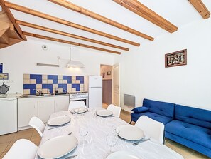 A dining area with a table set for four, a kitchen with a stove and refrigerator, a blue sofa, and wooden beams on the ceiling. - Maison Clos De Kermilienne by Interhome (Erdeven)