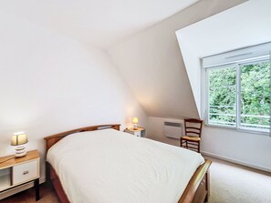 Een slaapkamer met een bed, nachtkastje, lamp, stoel en een raam met uitzicht op groen.