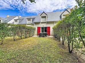 Ein zweistöckiges Haus mit roter Tür, einer kleinen Terrasse mit Tisch und Stühlen sowie einem Garten mit geschnittenen Büschen.