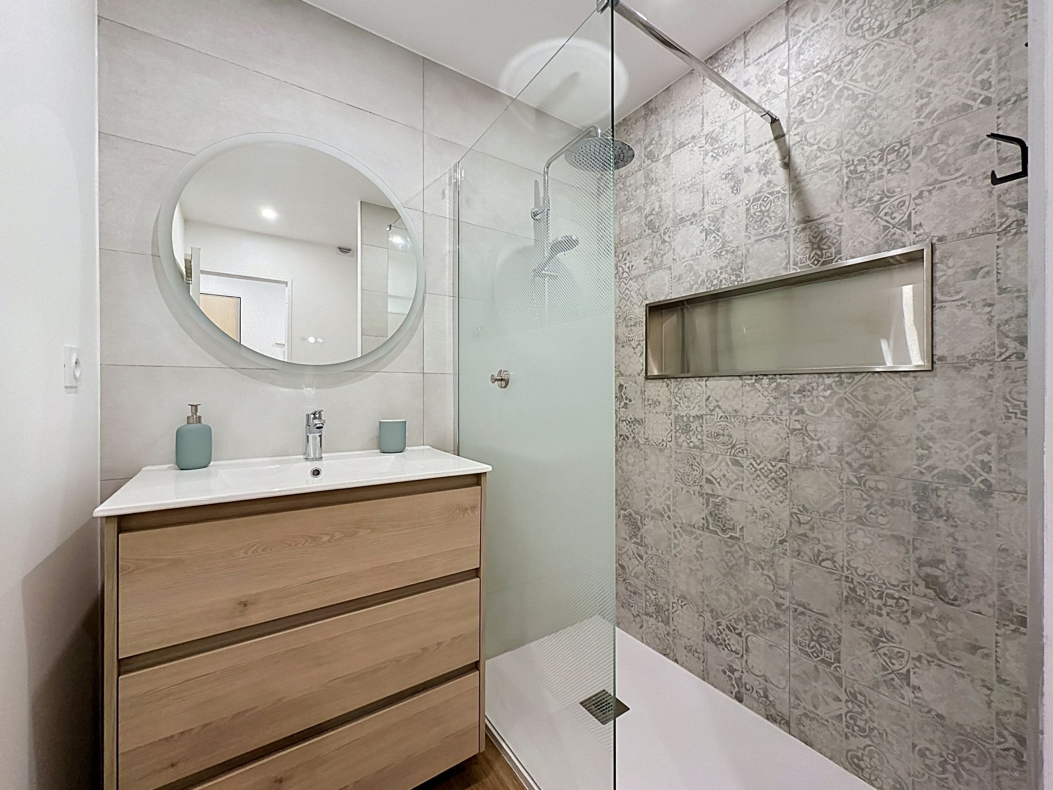 A modern bathroom with a round mirror, a wooden vanity, a glass shower enclosure, and patterned tiles.