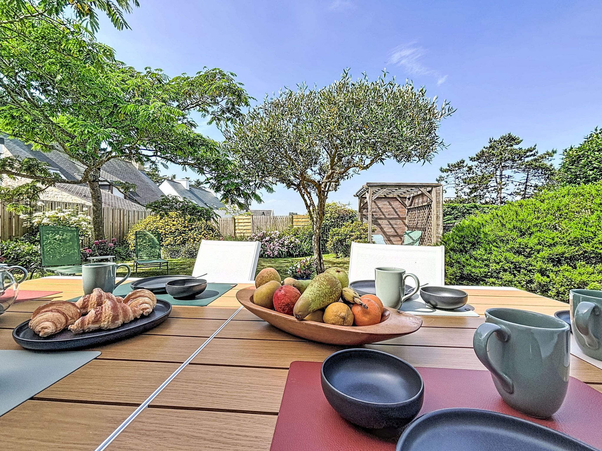 Un área de comedor al aire libre con una mesa de madera dispuesta para el desayuno, con croissants, frutas y un tazón de nueces.