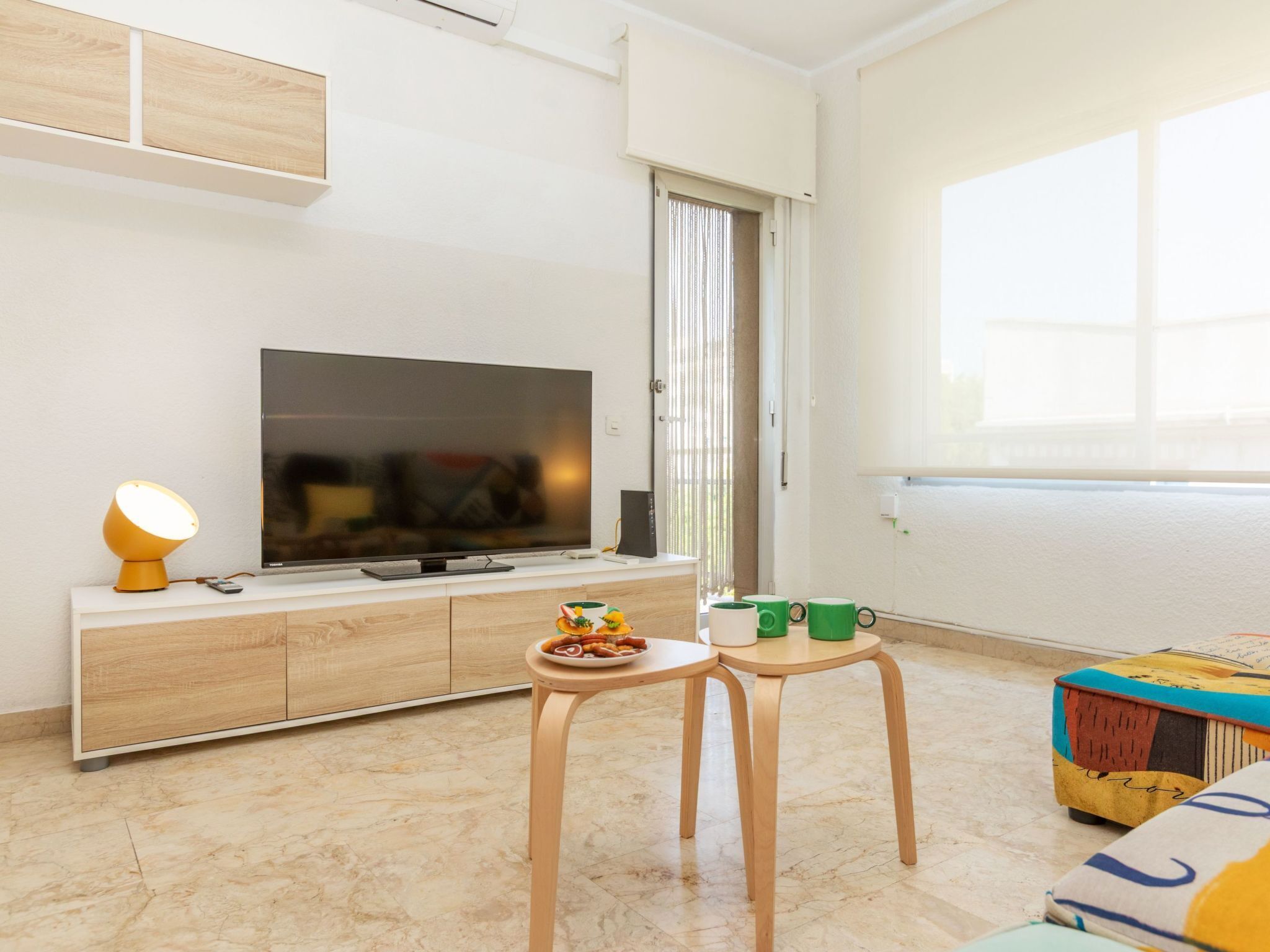 A modern living room with a flat-screen TV, wooden cabinet, two small tables, and a colorful ottoman.