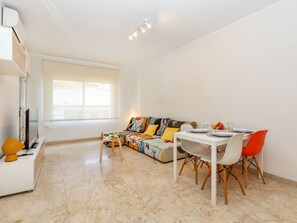 Ein modernes Wohnzimmer mit einer Couch, einem Esstisch und Stühlen. Ein Fernseher und ein kleiner Tisch mit Gegenständen darauf sind ebenfalls vorhanden.
