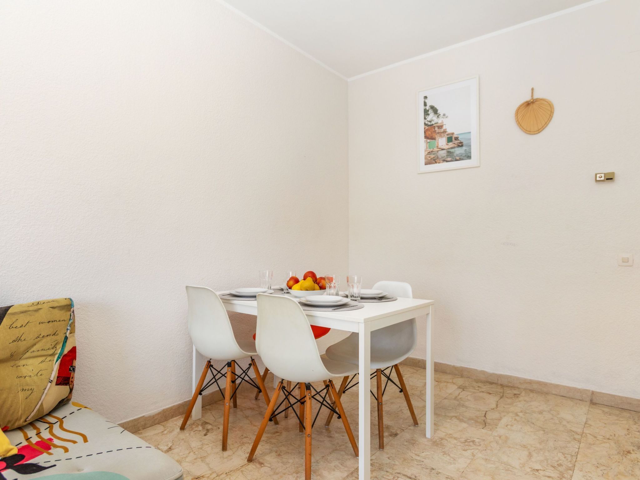 A dining area with a white table set for four, surrounded by white chairs with wooden legs. A bowl of fruit and glasses are on the table.