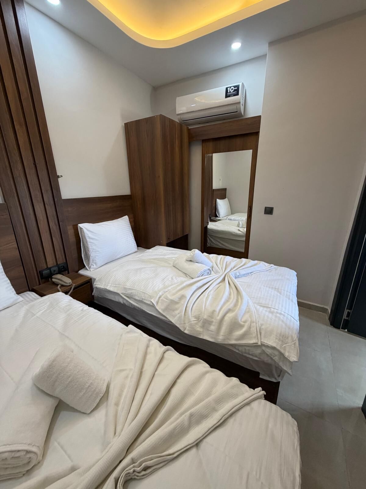 Camera d'hotel con due letti, una testiera in legno, un comodino con il telefono e un condizionatore d'aria.