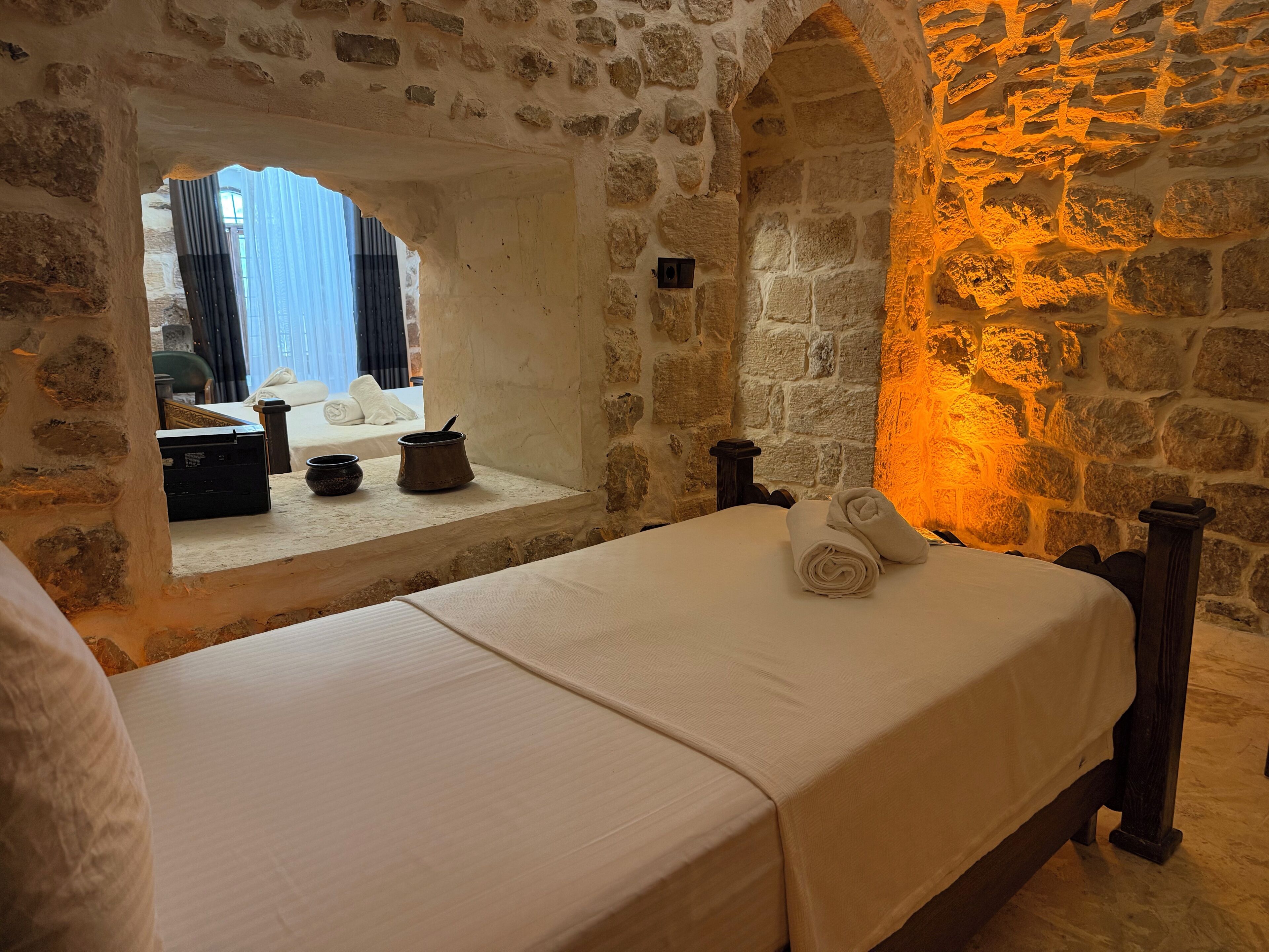 Una habitación con paredes de piedra, una cama, un ventanal y una pequeña mesa con una olla y un cuenco.