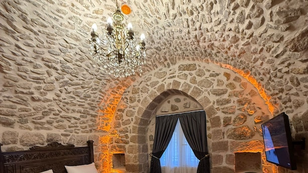 A stone-walled bedroom with a bed, a chandelier, a TV, and a chair. - Erkar Konağı (Mardin)