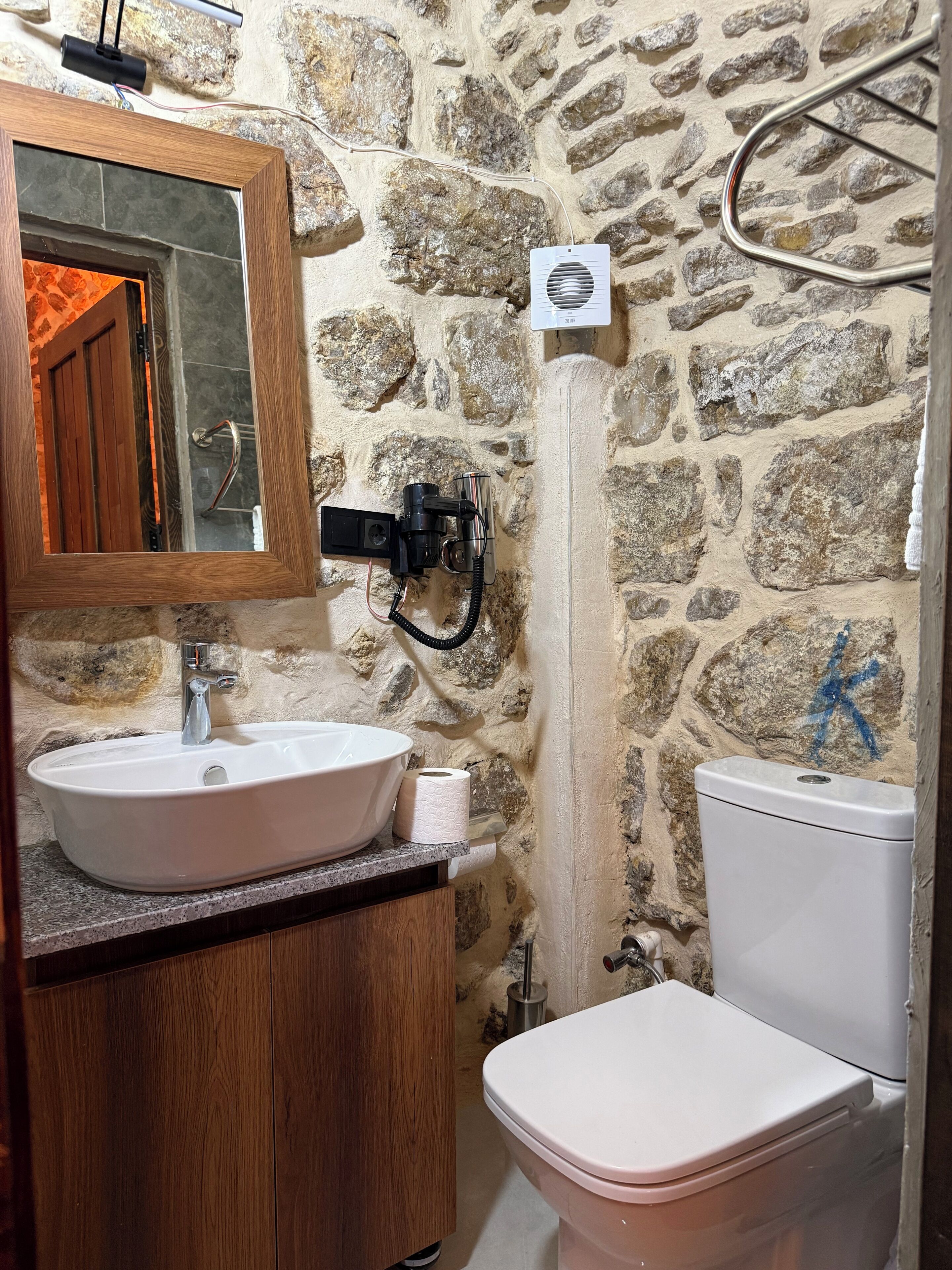 Un baño con pared de piedra, tocador de madera, lavabo blanco y un inodoro.