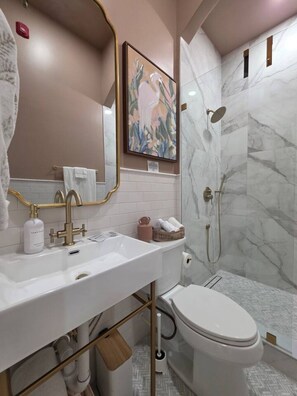 A modern bathroom with a white sink, gold fixtures, a shower with marble walls, and a framed artwork on the wall. - Luxury Queen Studio - Solara at The Marle Boutique Hotel-Near JHU + Downtown (Baltimore)