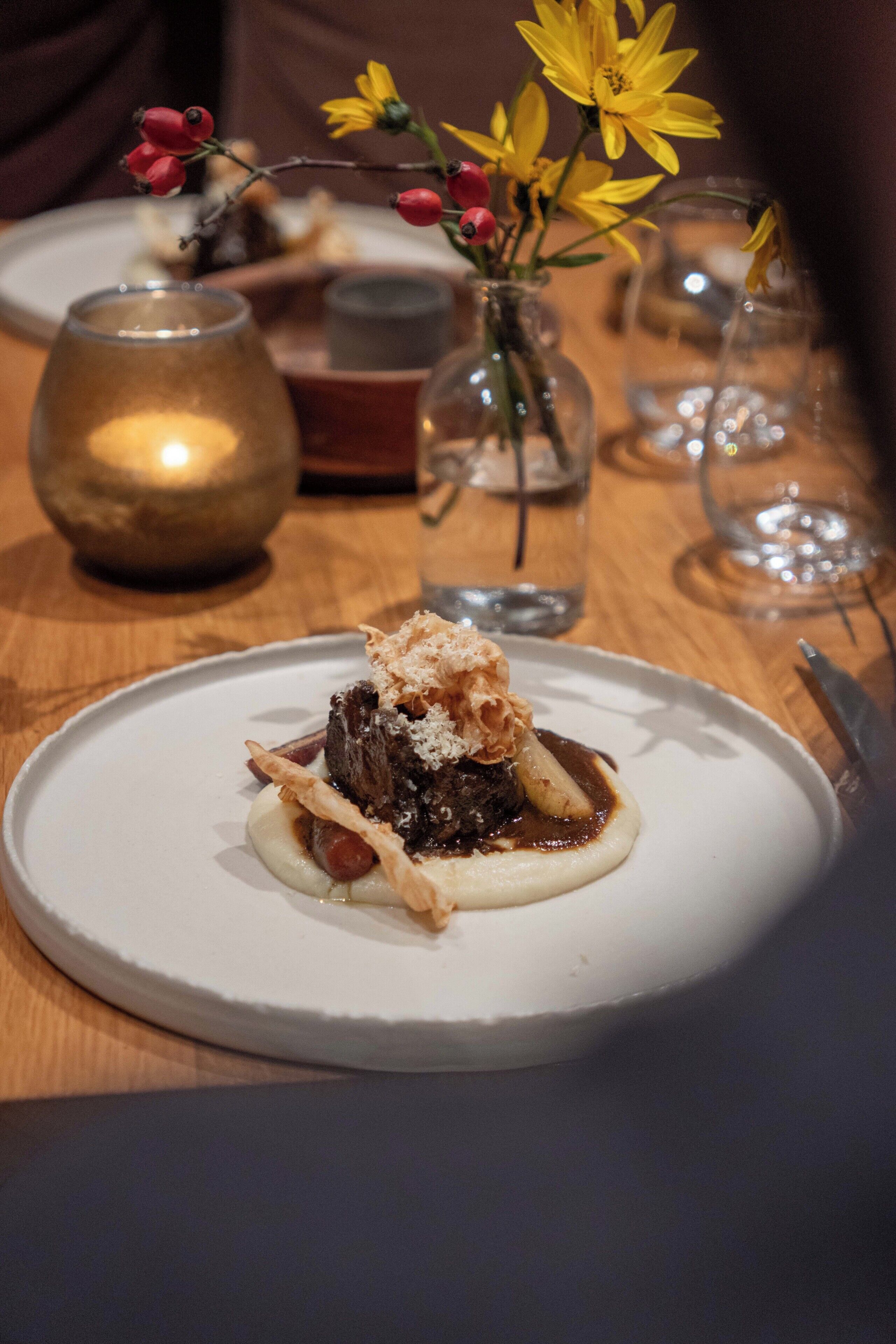 Un postre sobre un plato blanco, con una vela y un jarrón de flores amarillas al fondo.