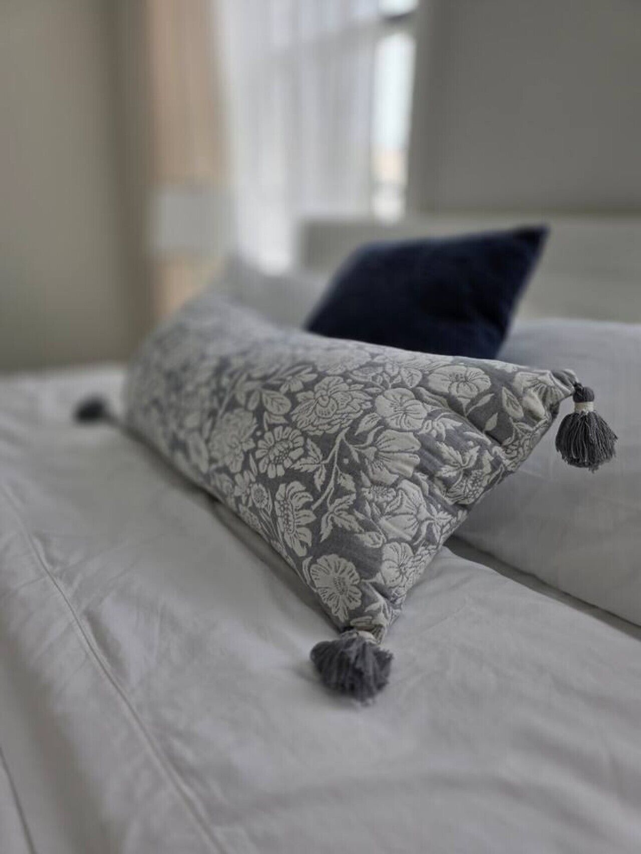 A bed with a floral-patterned pillow and a tassel, a dark blue pillow, and a window with blinds in the background.