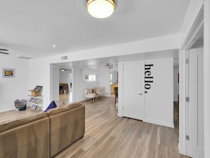 Ein modernes Interieur mit Holzboden, einem beigefarbenen Sofa, einer weißen Tür mit dem Schriftzug 'hello' und einem Flur, der zu einem Essbereich führt.