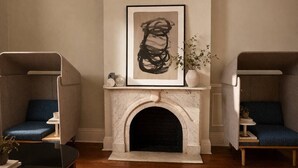 A room with a fireplace, two armchairs, a small table, and a framed artwork above the fireplace.