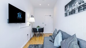 Ein modernes Wohnzimmer mit einem Flachbildfernseher, einem Essbereich mit Tisch und Stühlen, einem grauen Sofa und einem Stadtbildgemälde.
