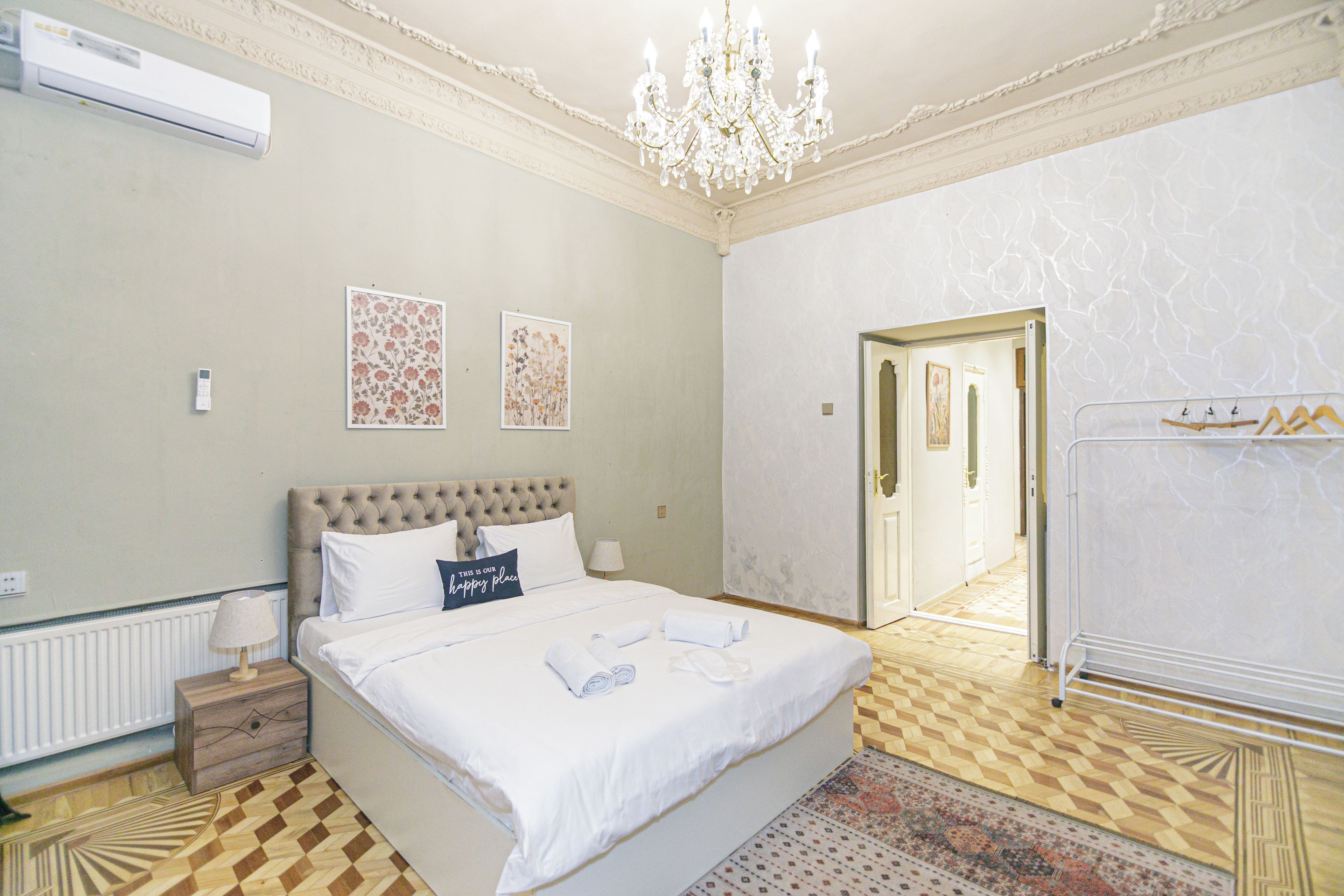 A hotel room with a large bed, a chandelier, a radiator, and a door leading to another room.